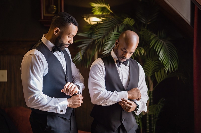 Groom and best man in tuxedos adjusting their watches.