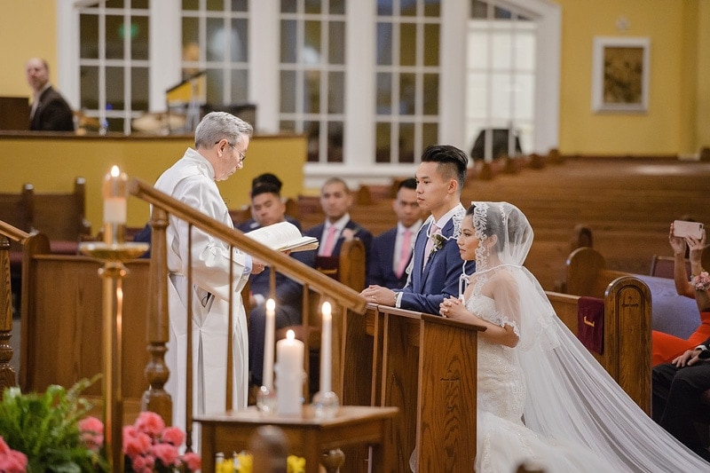 A bride and groom stand in front of a priest during their wedding ceremony, captured by skilled wedding photographers.