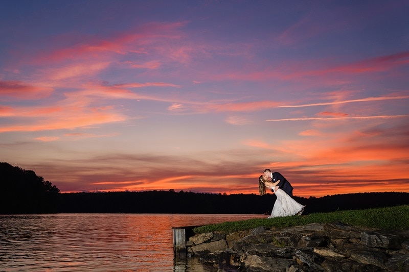 A bride and groom kiss in front of the Bella Collina Mansion at sunset, with a lake as their picturesque backdrop.