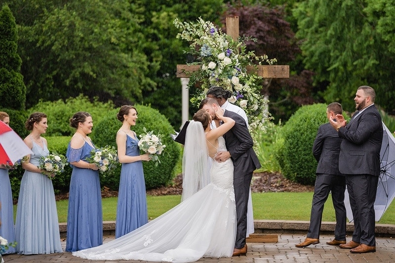 A wedding photographer captures the poignant moment when a bride and groom share a heartfelt kiss in front of their ecstatic wedding party.