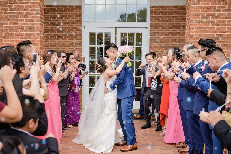 A bride and groom captured in a memorable moment, sharing a kiss against the backdrop of a charming brick building, beautifully portrayed by talented wedding photographers. The air filled with whimsical bubbles adds an enchanting