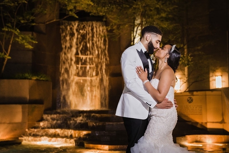 A bride and groom kiss in front of a fountain at their wedding reception.