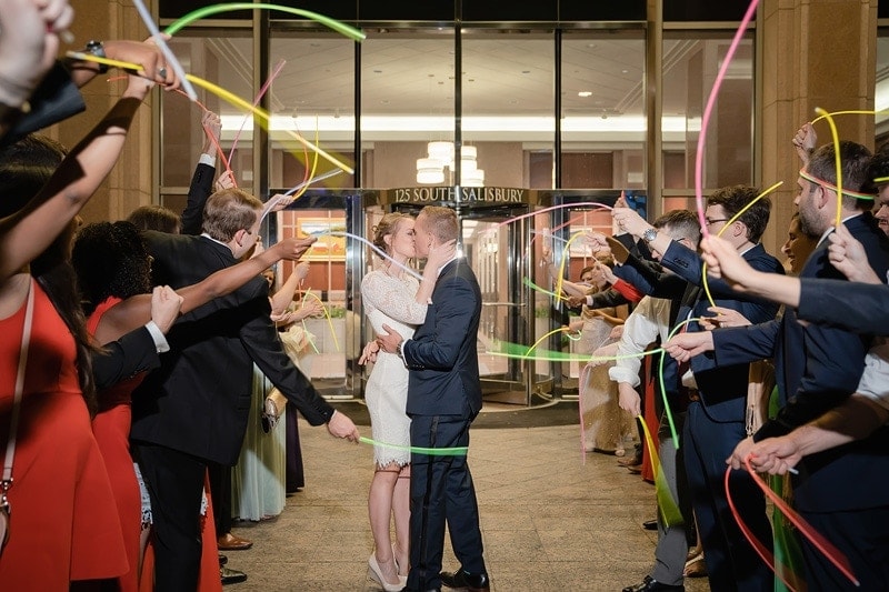 A newly married couple sharing a romantic kiss in front of a vibrant building adorned with colorful wands at their wedding.
