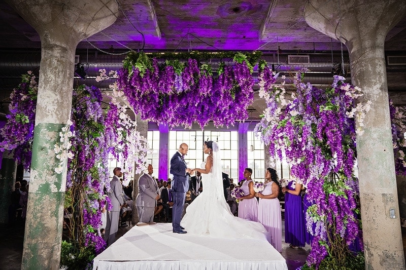 A gallery-style wedding ceremony with purple flowers hanging from the ceiling.