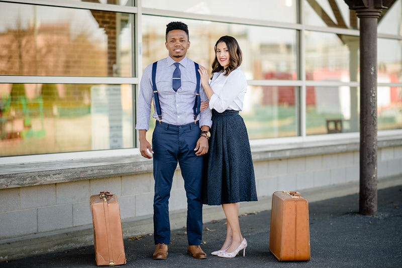 A young couple posing with suitcases in front of a train station for their engagement photos.