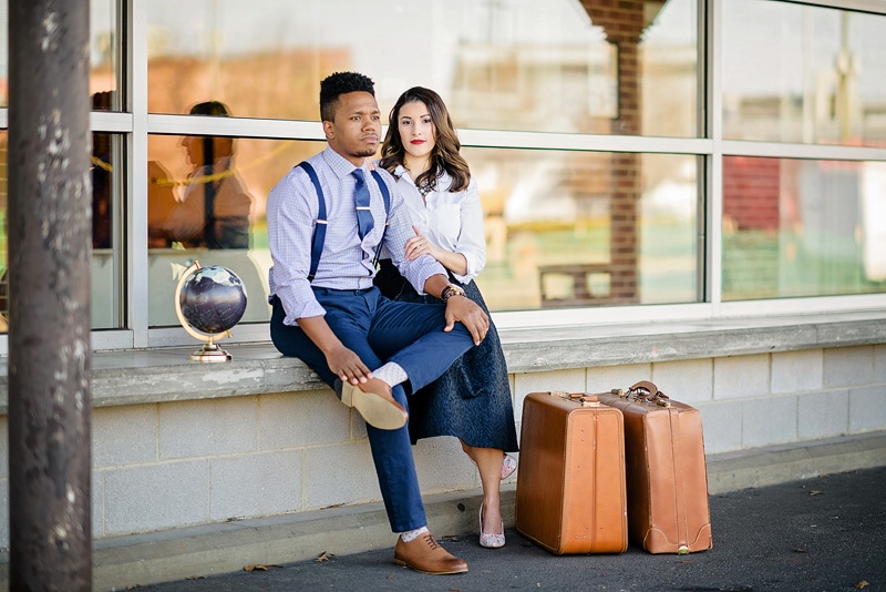 A man and woman, capturing their love story, sitting on a bench with suitcases during an engagement photoshoot by a professional engagement photographer.