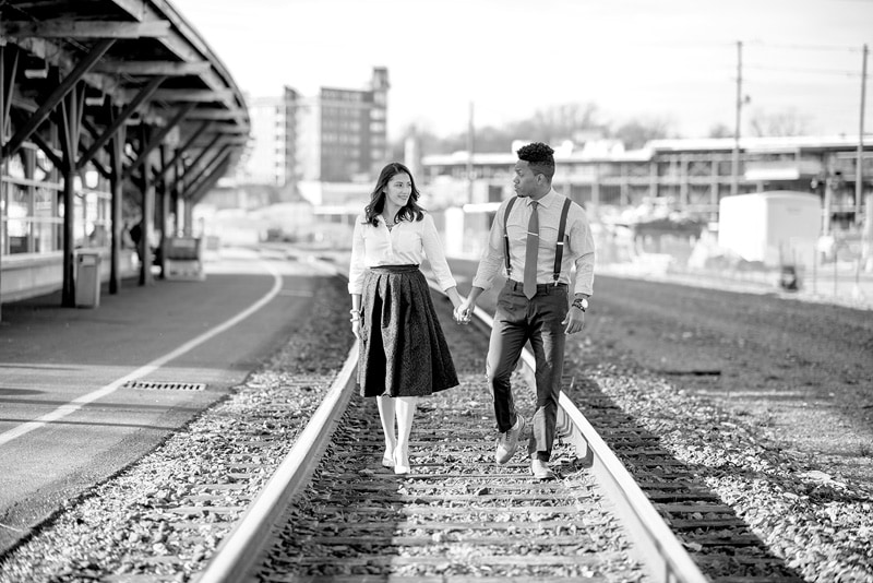 A black and white engagement photo of a couple walking on railroad tracks.