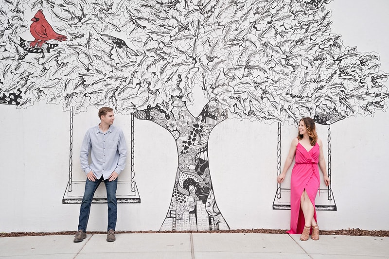 An engaged couple standing in front of a tree painted on a wall, captured beautifully by their engagement photographer.