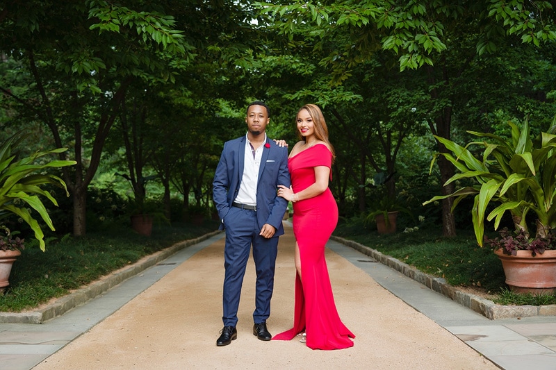 An engaged man and woman in a red dress standing on a path, captured beautifully by an engagement photographer.