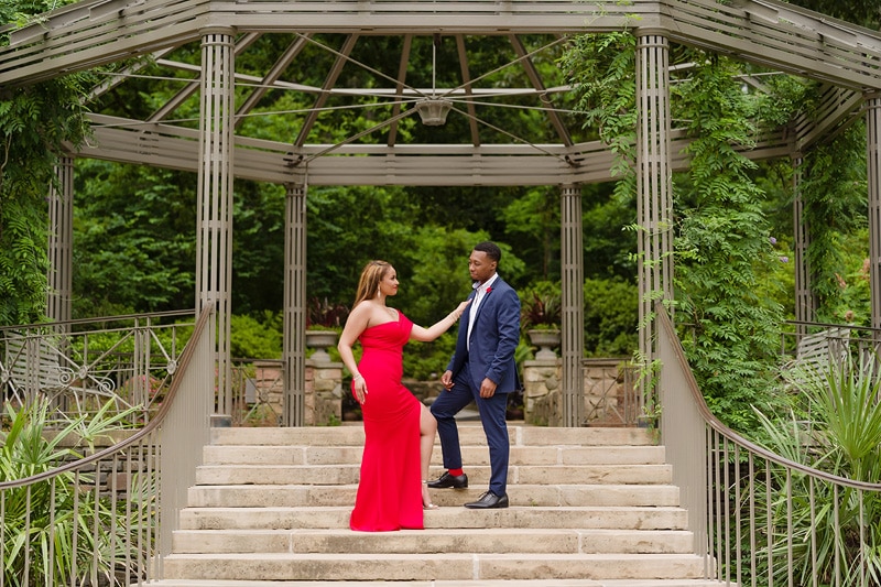 An engagement photographer capturing a couple in a red dress standing in front of a gazebo for their engagement photos.
