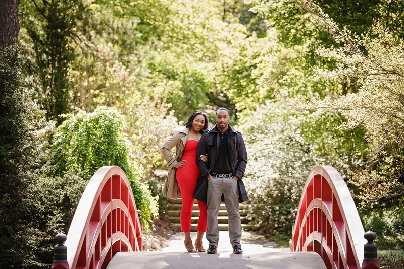 An engaged couple standing on a red bridge in a garden, captured by an engagement photographer.