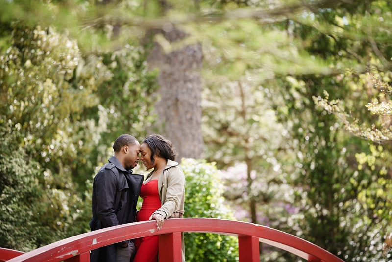 A couple embraces on a red bridge in a garden as their engagement photographer captures stunning engagement photos.