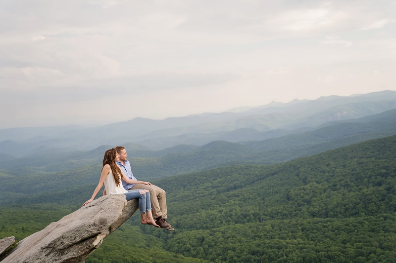 An engagement photographer captures a couple sitting on top of a cliff, creating stunning engagement photos with the breathtaking mountain view as their backdrop.