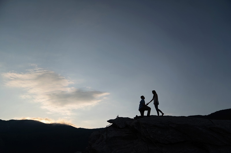 A man and woman capture their engagement moment on top of a rock at sunset with the help of an engagement photographer.