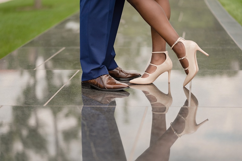 An engagement photographer captures a stunning image of a man and woman in high-heeled shoes standing on a reflective surface during their engagement photoshoot.