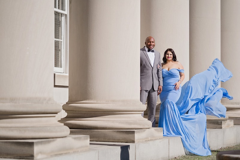 An engaged couple poses in front of columns in a blue dress for their engagement photos.