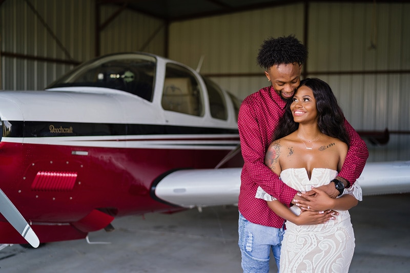 An engagement photoshoot capturing a couple embracing in front of an airplane hangar.