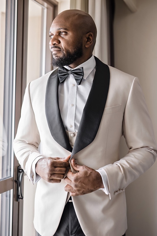 A black man in a white tuxedo and bow tie, getting ready for his wedding at The Distillery.