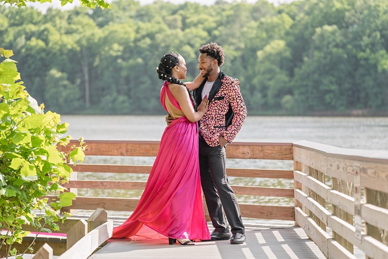 An engaged couple in a pink dress standing on a dock near a lake, captured by an engagement photographer.