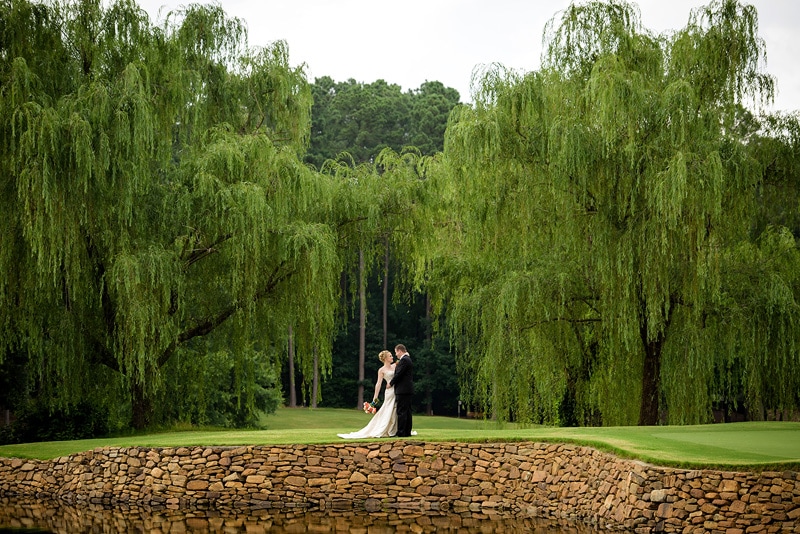 Wedding photographers capturing a bride and groom amidst serene willow trees by a pond.