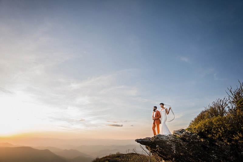 Wedding photographers capture a breathtaking moment as a bride and groom exchange vows on top of a cliff during a stunning sunset.