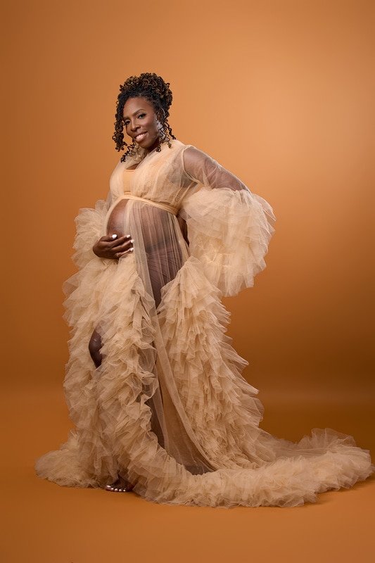A pregnant woman in a beige dress posing for maternity photos on an orange background.