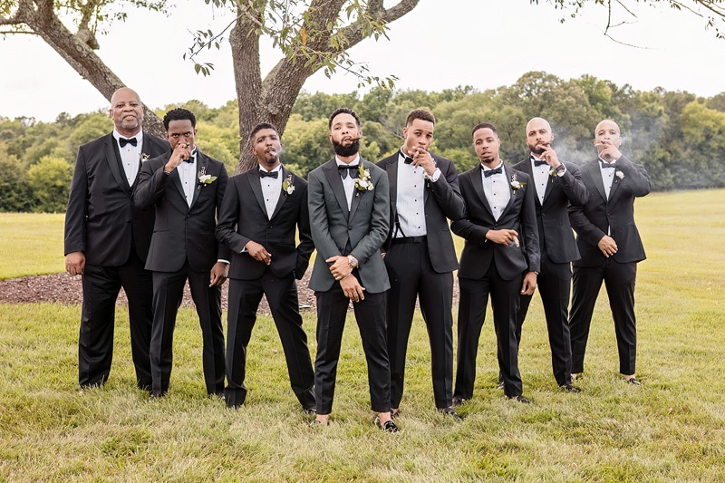 A group of groomsmen in tuxedos standing in front of a tree at a Board and Batten Wedding.