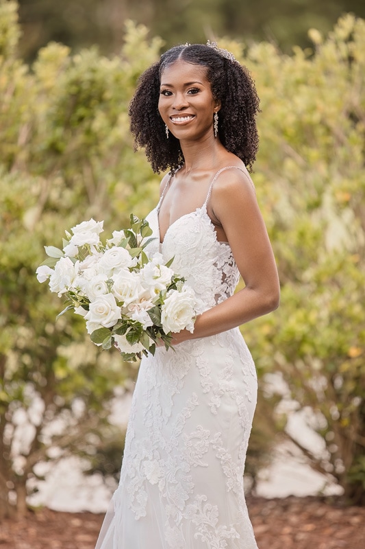 A black woman holding a bouquet of flowers in front of bushes at a Board and Batten wedding.