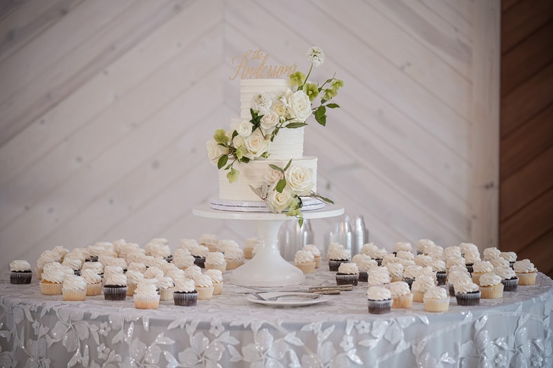 A white wedding cake and cupcakes are displayed on a table at a Board & Batten Events Wedding.