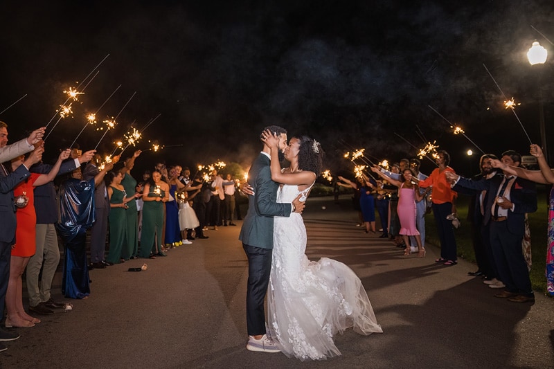 A bride and groom sharing a kiss in front of a crowd of sparklers at a Board & Batten Wedding.