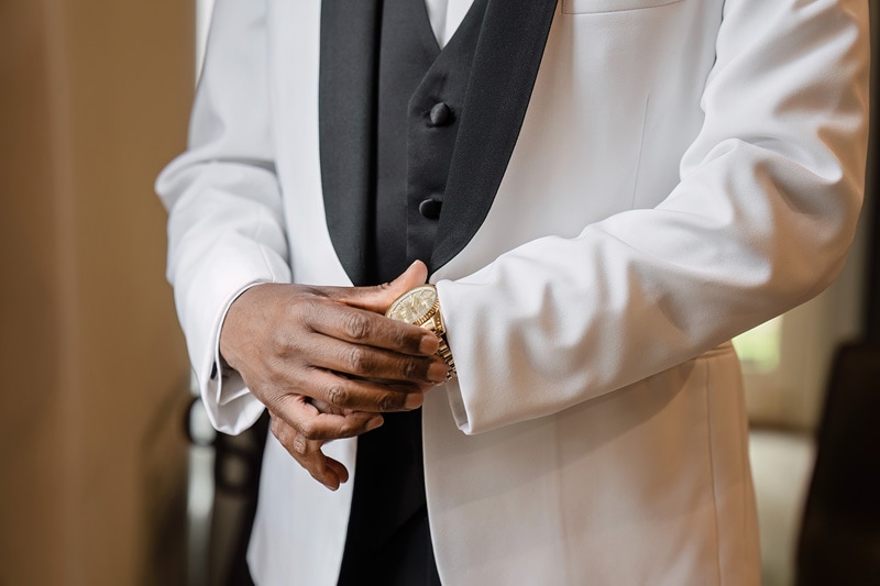 A man at the Pinehill Pavilion Wedding is putting on his watch in a white tuxedo.