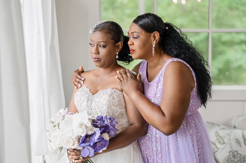 The bride and maid of honor hugging each other in front of a window at the Pinehill Pavilion wedding venue.