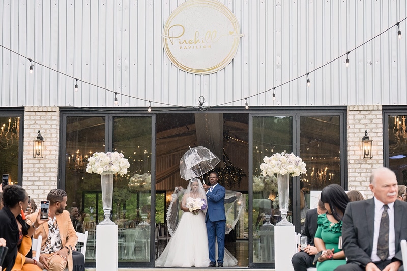 A Pinehill Pavilion wedding featuring the bride gracefully walking down the aisle with an umbrella, adding a touch of elegance to their special day.