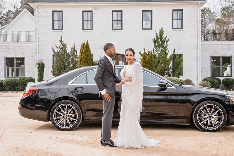 A man and woman in a suit and white dress standing at The Bradford wedding venue in front of a black car.