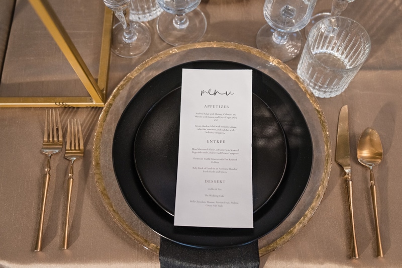 A table setting with a black plate and gold forks at The Bradford Wedding Venue.