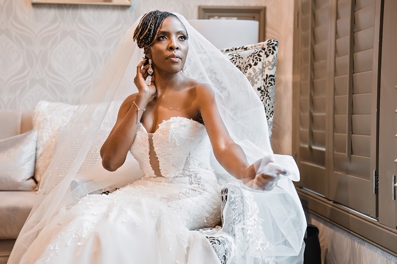 A bride in a white wedding dress and veil is seated at the Crystal Ballroom Charlotte, looking to the side while touching her earring.
