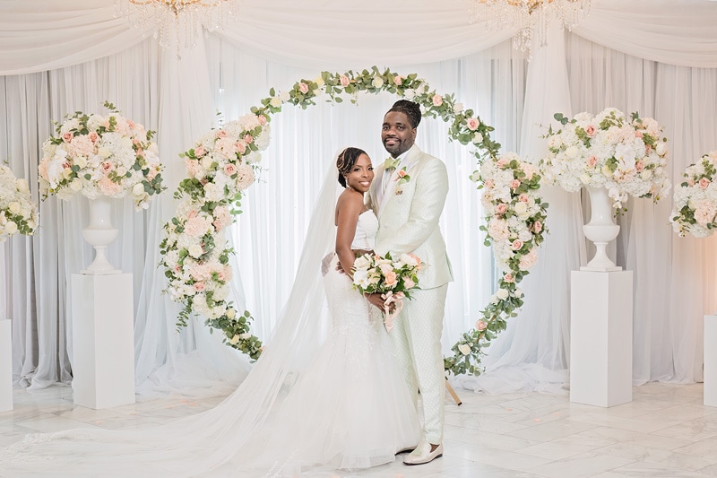 A bride and groom are posing for a photograph in the Crystal Ballroom Charlotte, an elegant wedding venue adorned with floral arrangements and drapery.