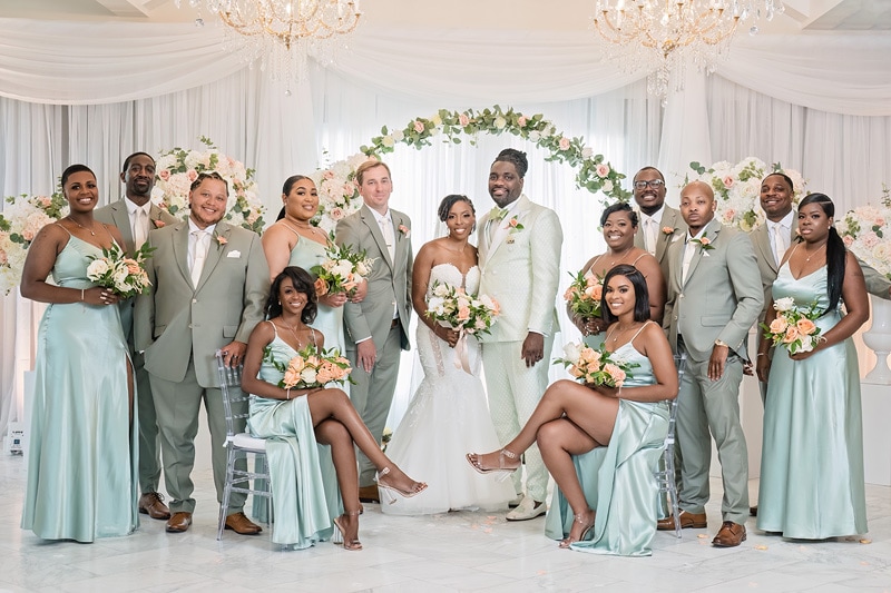 A wedding party, including the bride and groom, bridesmaids, and groomsmen, posing for a photo in the Crystal Ballroom Charlotte, a well-decorated venue with floral arrangements and ch