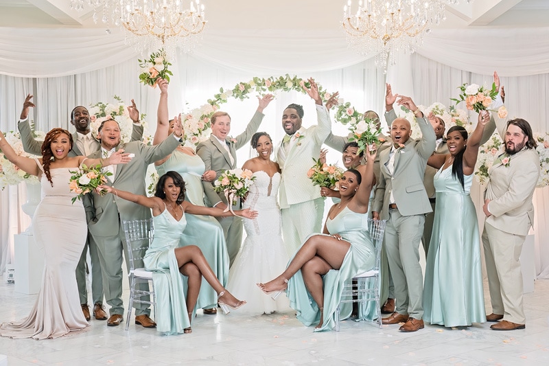 A joyful wedding party celebrating in the Crystal Ballroom Charlotte, with the bride and groom at the center surrounded by bridesmaids and groomsmen in coordinating attire, all cheering and posing in an elegantly