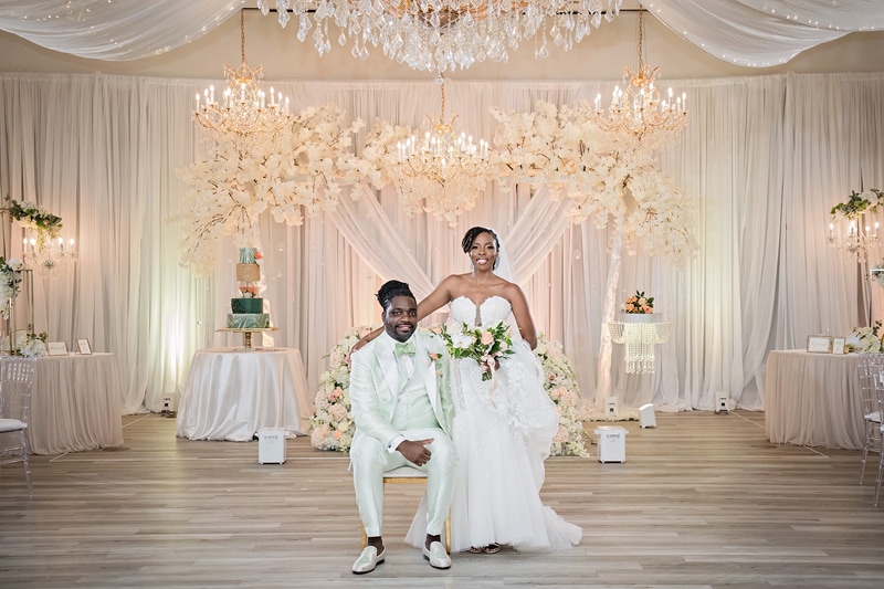 A couple in wedding attire posing with smiles in the Crystal Ballroom Charlotte, elegantly decorated with floral arrangements and a chandelier.