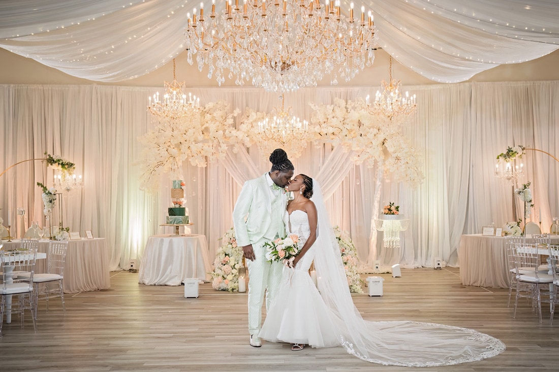 A bride and groom stand close to each other beneath an elaborate chandelier, in the beautifully decorated Crystal Ballroom Charlotte with elegant floral arrangements and a tiered wedding cake.