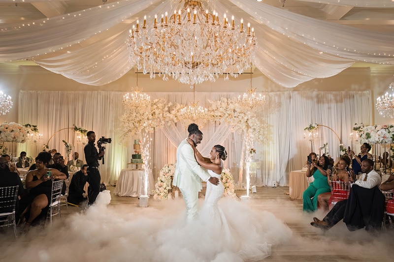A bride and groom share a dance surrounded by guests in the elegantly decorated Crystal Ballroom in Charlotte with draped ceilings, a chandelier, and artificial fog on the floor.