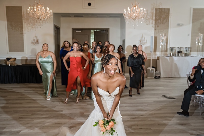 A bride in a white dress joyfully races ahead while her bridesmaids, dressed in various colors, trail behind her in the brightly lit Crystal Ballroom Charlotte.