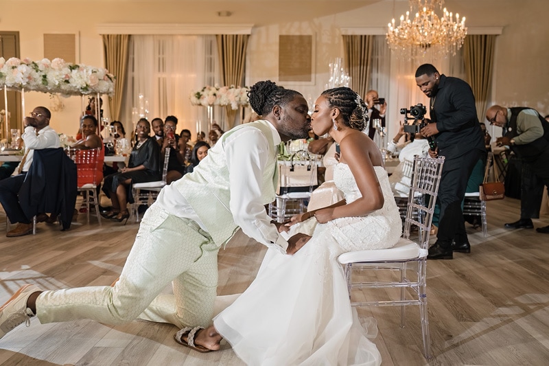 A newlywed couple shares a kiss while the groom is knelt on one knee in the Crystal Ballroom Charlotte, as guests watch and a photographer captures the moment during a wedding reception.