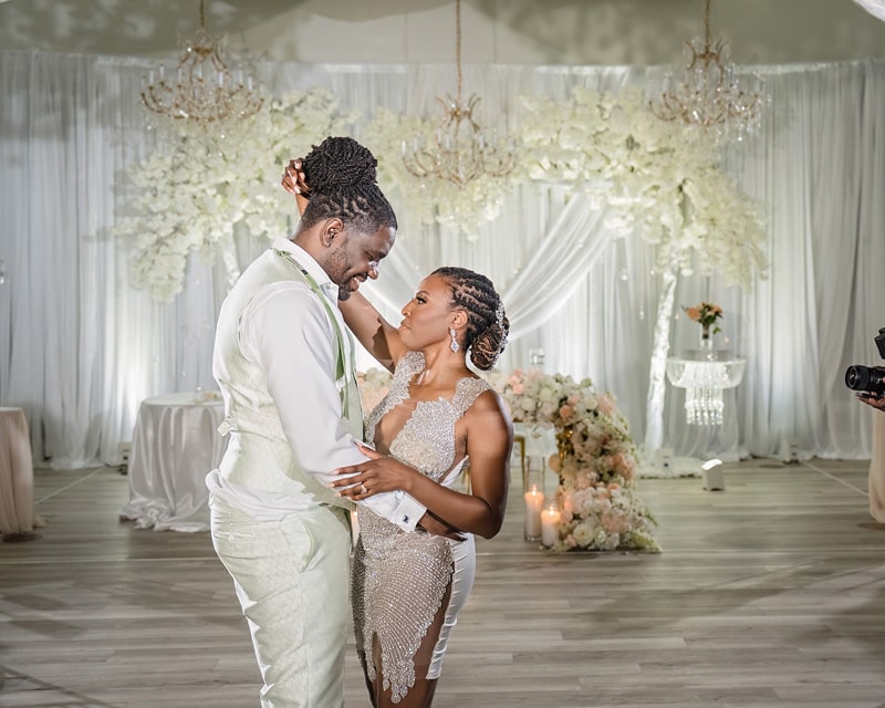 A bride and groom share a dance on their wedding day in the Crystal Ballroom Charlotte, elegantly decorated with chandeliers and floral arrangements, while a photographer captures the moment.