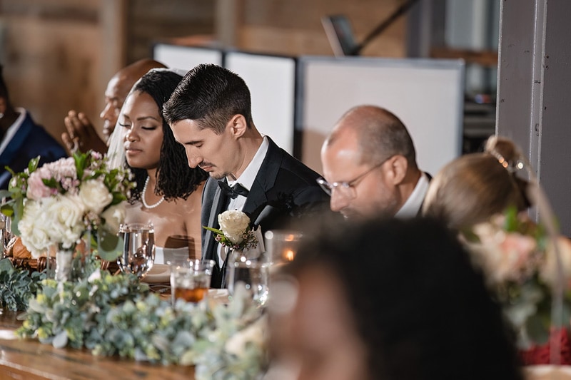 A groom in a black tuxedo thoughtfully listens at The Rickhouse wedding table adorned with flowers, surrounded by elegantly dressed guests in a warmly lit venue.