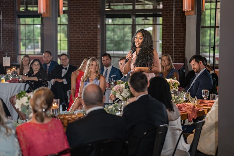 A wedding guest delivers an emotional speech at The Rickhouse wedding reception, holding the room's attention amidst elegant table settings and an atmosphere of warm, ambient lighting.