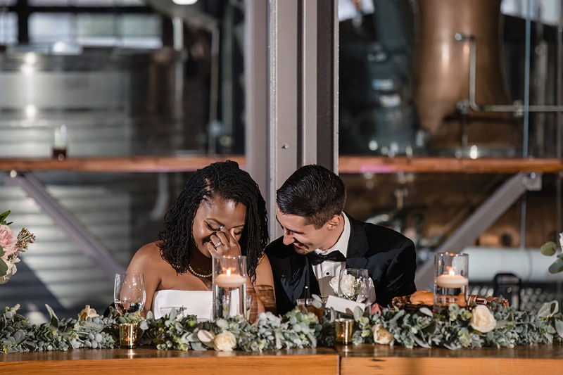 A couple shares an intimate, joyous moment at a candlelit table adorned with flowers, their laughter and affection shining amidst an elegant, industrial-chic setting at The Rickhouse wedding reception.
