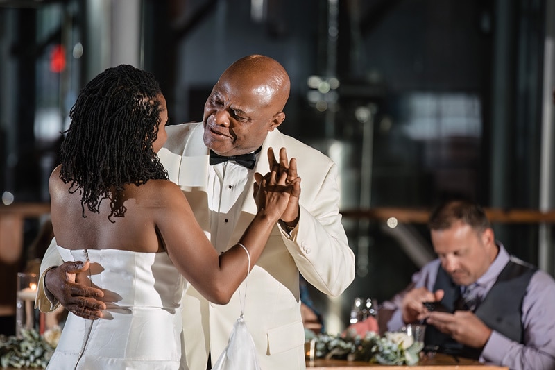 A couple dances intimately in The Rickhouse wedding attire, sharing a moment of connection, while a guest in the background checks his phone, surrounded by elegant table settings and soft candlelight.