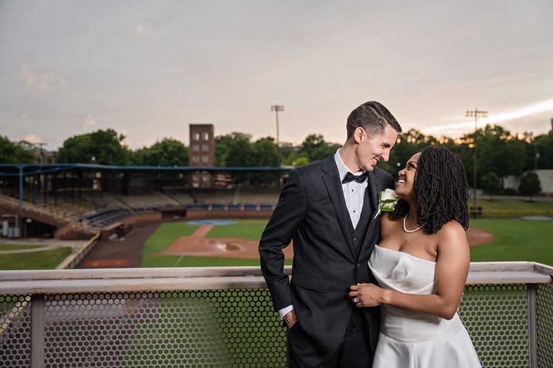 A couple in formal wedding attire shares a tender moment with a serene smile, standing on a balcony of The Rickhouse during their wedding reception, with a baseball stadium under the enchanting glow of a dus
